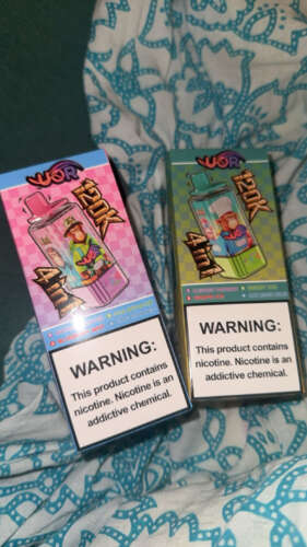 Two vape product boxes with vibrant designs, listing flavors like Fuji Apple Ice and Blueberry Raspberry, on a patterned fabric background