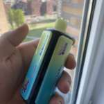 A hand holding a small blue and light-green vape device with a gradient color scheme, black mouthpiece, and colorful labels, near a window with a cityscape in the background.