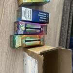 Several colorful “25X” energy shot boxes stacked next to a wooden table, with an open brown cardboard box at the bottom.