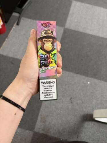 Hand die een kleurrijke vape-doos vasthoudt met het opschrift “30k puffs” met aardbei-kiwi-smaak; nicotinewaarschuwing onderaan.